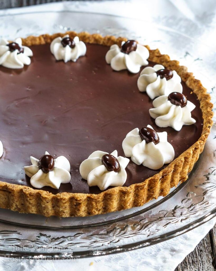 A round dark chocolate tart with piped stars of whipped topping garnished with chocolate-covered coffee beans sitting on a glass serving plate.