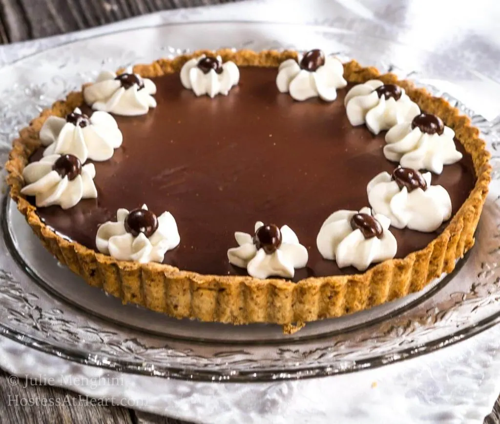 A Dark Chocolate Pecan Tart with piped stars of whipped topping and a chocolate-dipped coffee beans sitting on a glass plate over a white tablecloth.