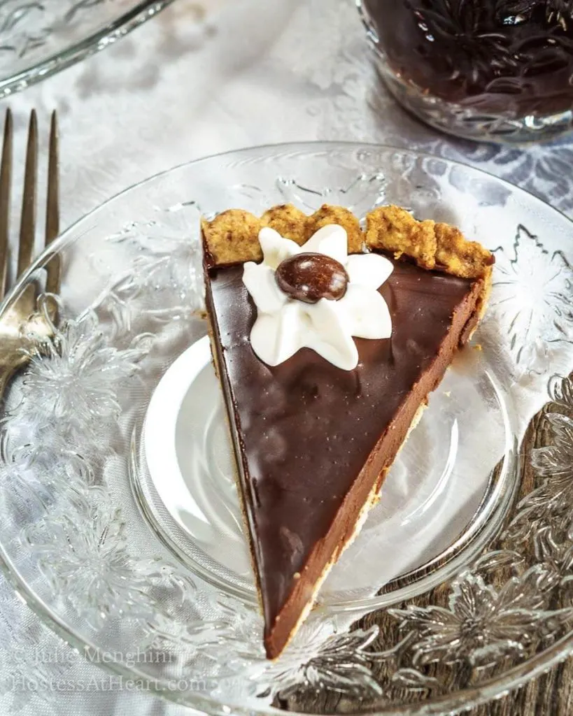 A slice of Dark Chocolate Pecan Tart with a piped star of whipped topping and a chocolate-dipped coffee bean sitting on a glass plate over a white tablecloth. A cup of coffee sits in the background.