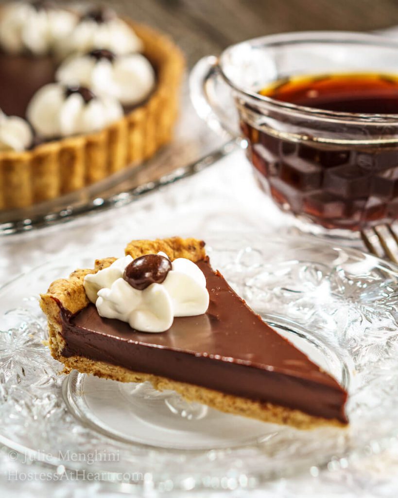 A slice of Dark Chocolate Pecan Tart with a piped star of whipped topping and a chocolate-dipped coffee bean sitting on a glass plate over a white tablecloth. A cup of coffee is in the background next to the whole tart.