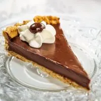 A slice of Dark Chocolate Pecan Tart with a piped star of whipped topping and a chocolate-dipped coffee bean sitting on a glass plate over a white tablecloth.
