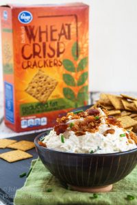 A bowl Cheese dip made with crab and bacon sits in a black bowl with crackers in the background. The box of crackers with next to the crackers.