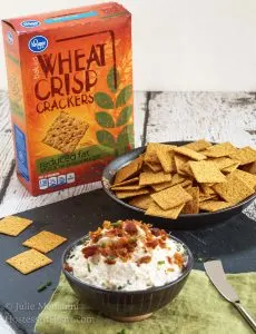 A bowl Cheese dip made with crab and bacon sits in a black bowl with crackers in the background. The box sits in the background.