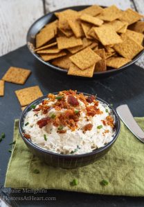 A bowl Cheese dip made with crab and bacon sits in a black bowl with crackers in the background.