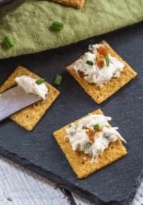 Three crackers topped with crab dip sitting on a slate tray next to a spreading knife.