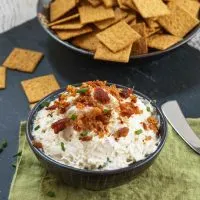 A bowl of Crab Dip with bacon sitting on a green napkin. A bowl of crackers sits in the background.
