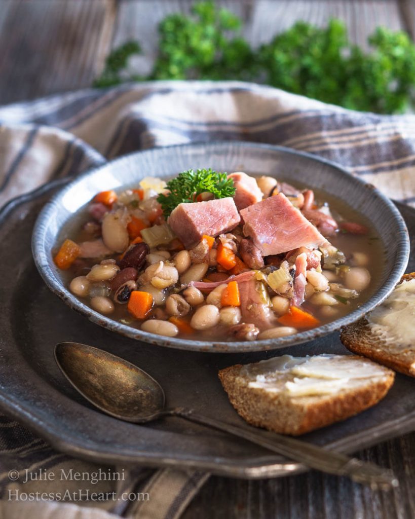 A bowl of Ham and bean soup garnished with big chunks of ham. Sliced bread and a spoon sits next to the bowl on a metal plate.