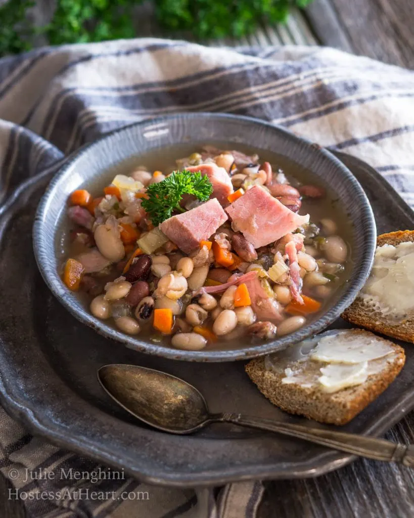 A bowl of Ham and bean soup garnished with big chunks of ham over a striped blue towel. Sliced bread and a spoon sits next to the bowl on a metal plate.