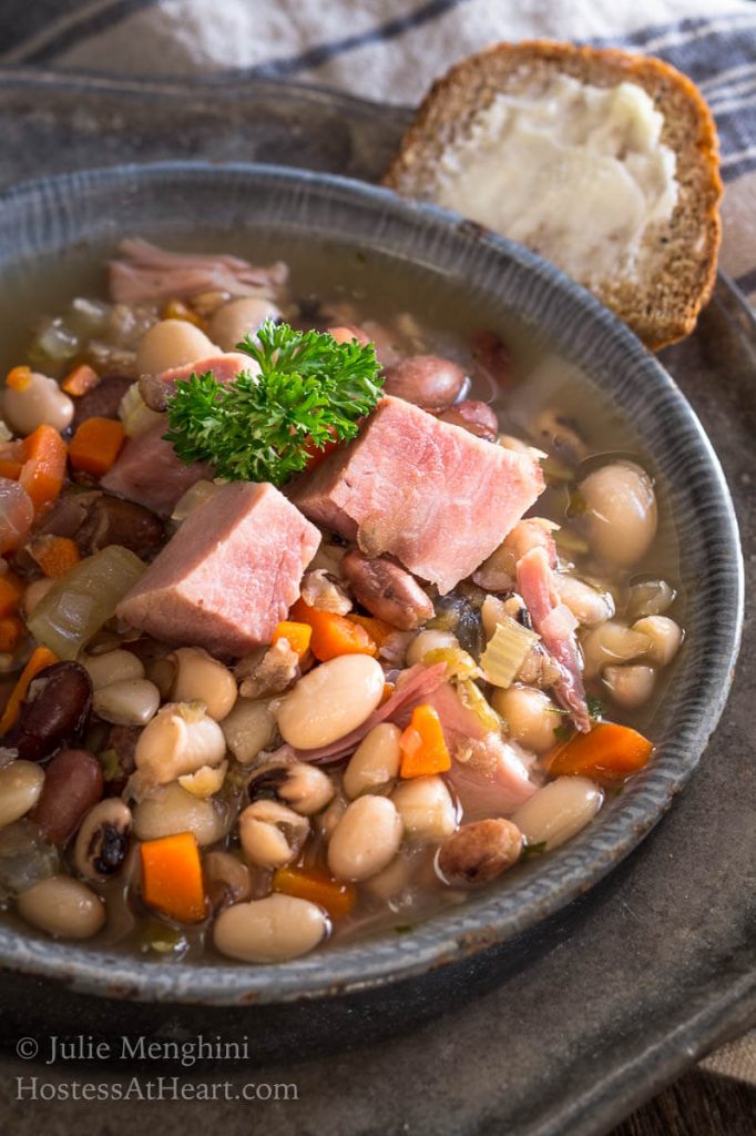 Top-down view of a bowl of Ham and bean soup garnished with big chunks of ham. Sliced bread and a spoon sits next to the bowl on a metal plate.