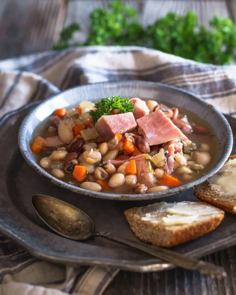 Blue granite bowl filled with Ham and Bean Soup containing both shredded ham and chunks of ham, carrots, and herbs. The bowl sits on a metal plate next to sliced bread and a spoon.  A striped napkin and fresh parsley sit in the background.