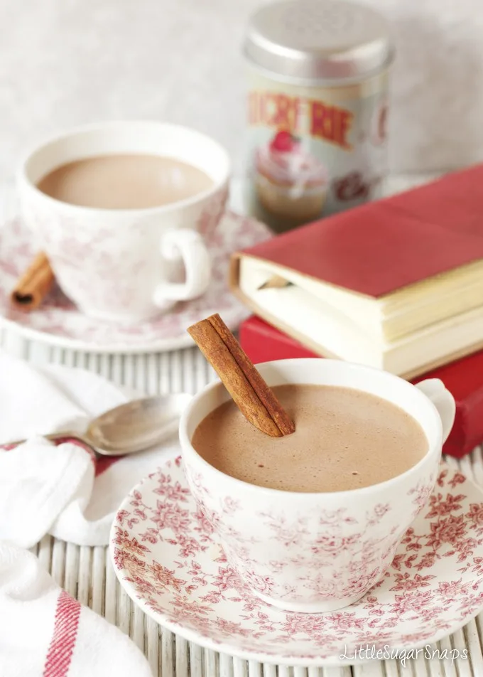 A cup of cinnamon hot chocolate in a patterned coffeecup with a cinnamon stick sitting in it in front of a stack of books and a second cup in the background.  