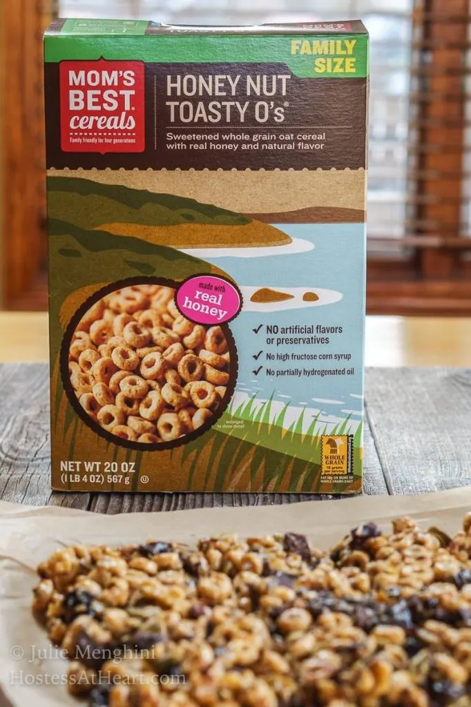 A healthy snack bar made from cereal, honey, and dark chocolate in front of a box of cereal.