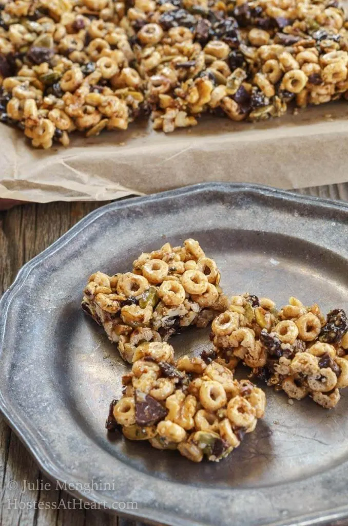 Three bars made from honey, whole-grain cereal, and chocolate chunks sitting on a metal plate in front of a pan of bars.