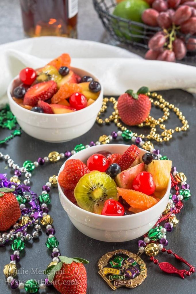 A two bowls fruit salad infused with Hurricane cocktail mix over gray slate and surrounded with Mardi Gras beads.