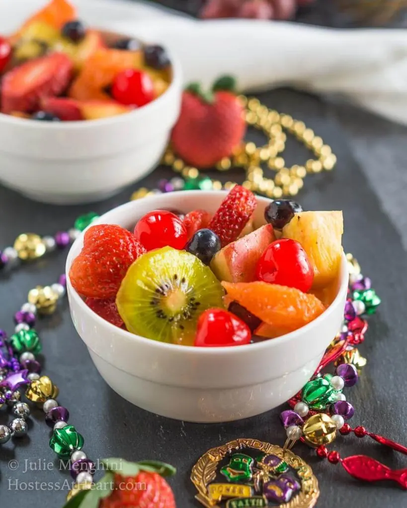 A two bowls fruit salad infused with Hurricane cocktail mix over gray slate and surrounded with Mardi Gras beads.