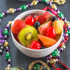 A bowl of Hurricane infused fresh fruit sitting on black slate and surrounded with Mardi Gras beads.