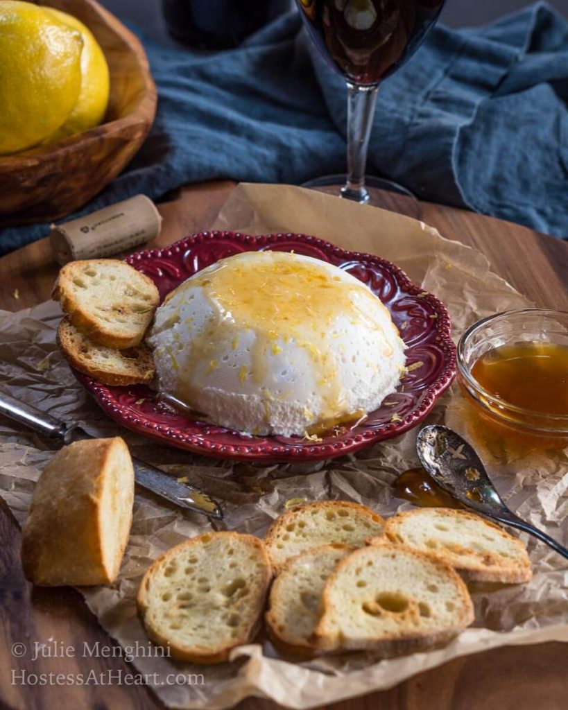 Red plate holds a bowl-shaped homemade ricotta drizzled with honey with slices of a baguette sitting in the front over a wooden cutting board.