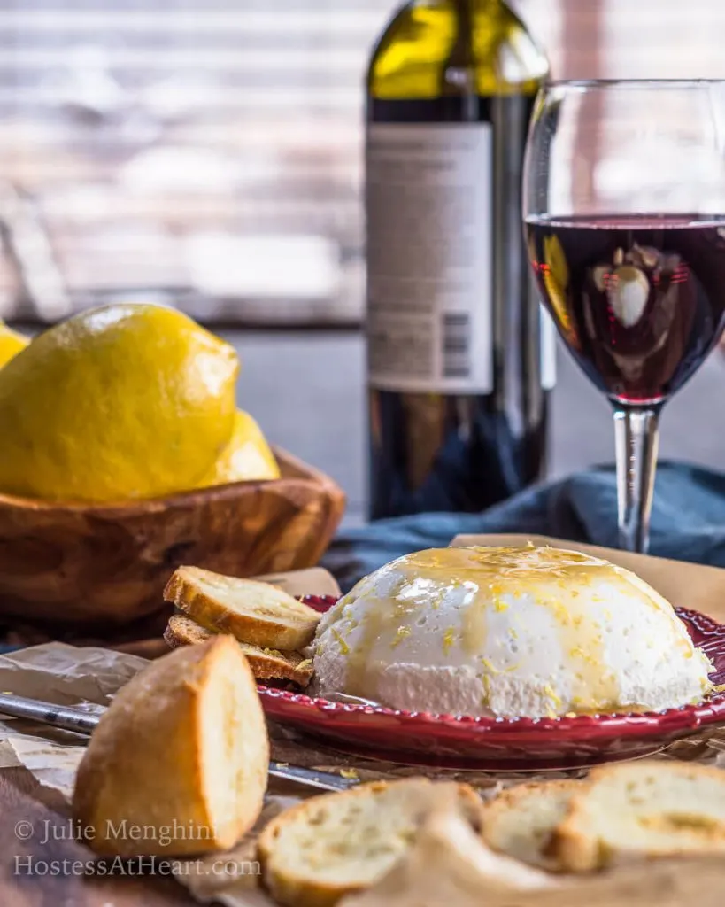 Side scene of homemade ricotta drizzled with honey sitting on a red plate in front of a bottle of a glass and bottle of red wine and fresh lemons.