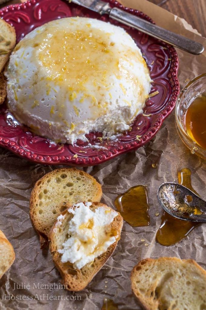 Homemade Lemon Ricotta sits on a red plate drizzled with honey over a piece of parchment paper. A sliced baguette sits in front spread with the ricotta and drizzled with honey.