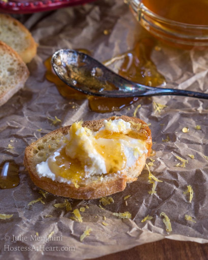 A baguette slice with a mound of fresh ricotta spread on top and then drizzled with honey. An antique spoon sits in the back over a piece of parchment paper.