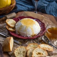 Red plate holds a bowl-shaped homemade ricotta drizzled with honey with slices of a baguette sitting in the front over a wooden cutting board.