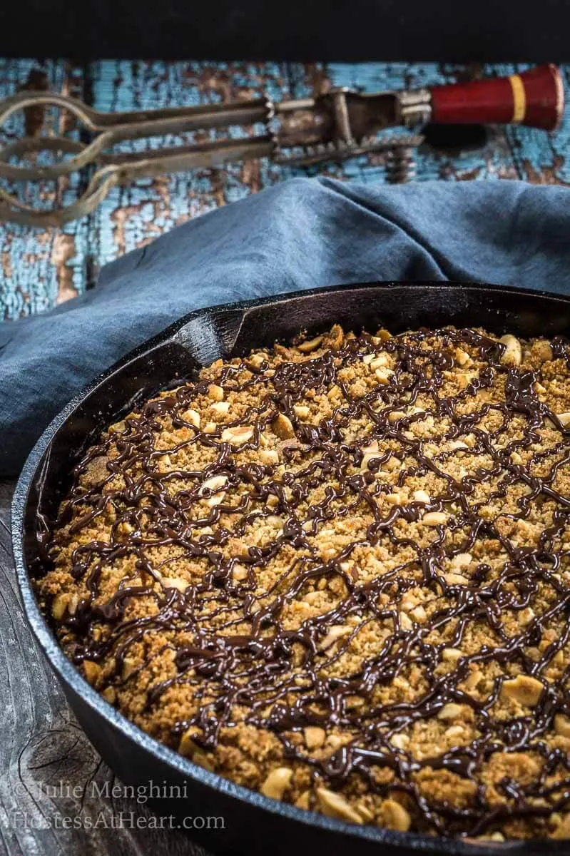 Top angle view of a peanut butter coffee cake in a cast-iron skillet drizzeled with Nutella and sitting on a blue napkin with an antique egg beater behind it 