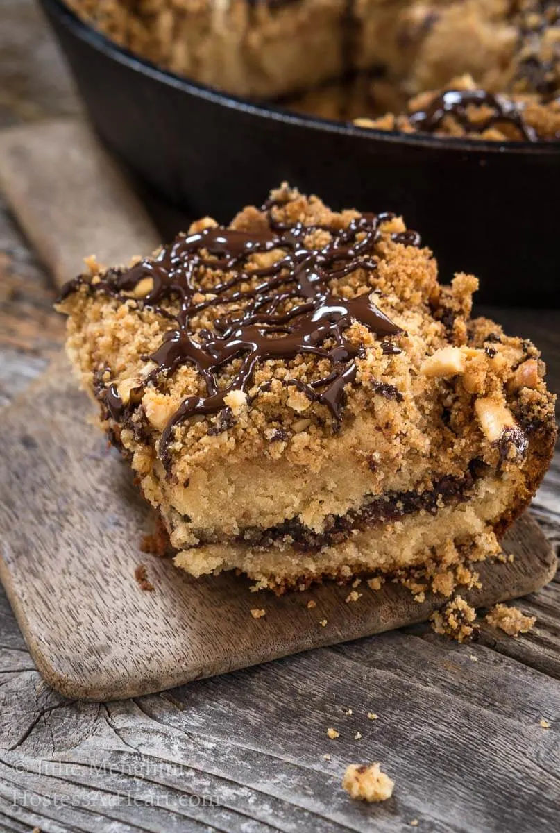 A piece of peanut butter coffee cake shows a nutella layer with a crumb topping drizzled with chocolate sitting on a wooden butter paddle