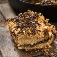 Piece of peanut butter coffee cake drizzled with chocolate is sitting on a wooden paddle. The slice shows a line of chocolate running through it. A cast-iron pan filled with the coffee cake sits in the background.