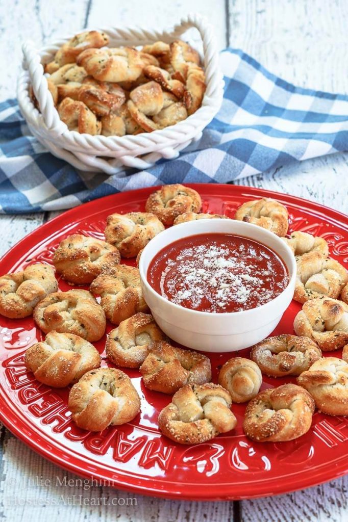 Pizza Knots sit on a bright red plate with a bowl of marinara in the center.  White basket filled with more pizza knots over a blue checked napkin in the background.