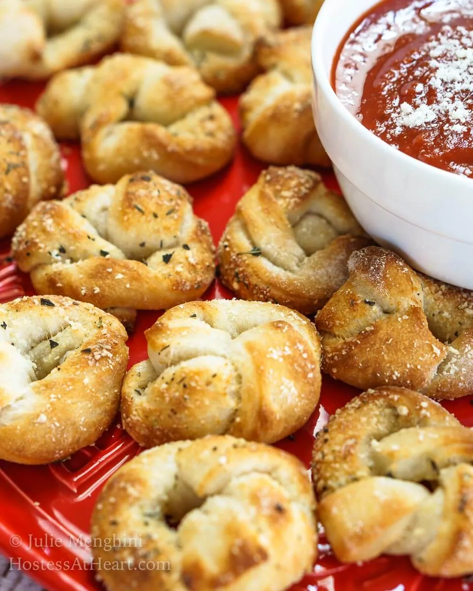 Close-up photo of a red plate topped with baked pizza knots and a white bowl containing pizza sauce that\'s dusted with grated cheese..