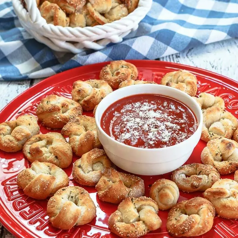 Red plate topped with baked pizza knots and a white bowl containing pizza sauce. A white basket of pizza rolls sit in the background over a blue checked napkin.