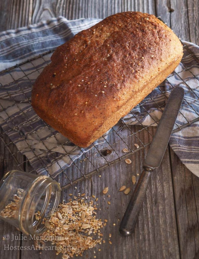 Top down view of a perfectly browned loaf of whole grain bread sprinkled with seeds and grains. The bread sits on a cooling rack over a blue striped napkin on a wooden backdrop. A small jar of the whole grain seed mix is topped over on it\'s side spilling onto the wooden backdrop.