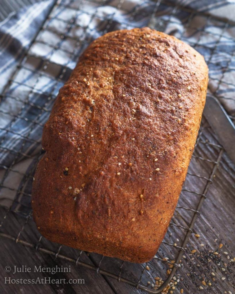 Top down view of a perfectly browned loaf of whole grain bread sprinkled with seeds and grains sitting at an angle over a cooling rack over a blue striped napkin on a wooden backdrop.