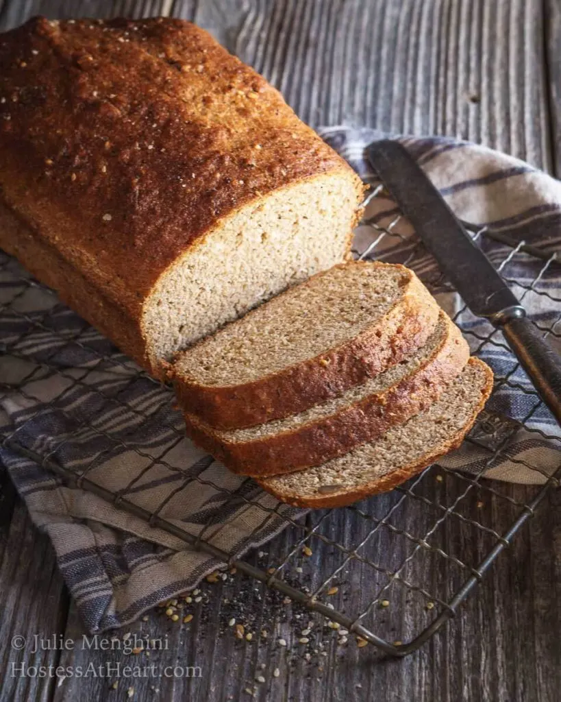 Side angle of a loaf of whole-grain bread with 3 slices cut from the front showing the soft crumb of this perfectly browned bread. The loaf sits on a cooling rack over a striped napkin and next to an antique butter knife.