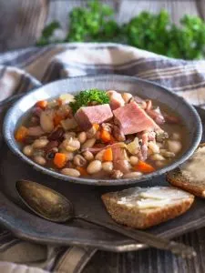 Blue granite bowl filled with Ham and Bean Soup containing both shredded ham and chunks of ham, carrots, and herbs. The bowl sits on a metal plate next to sliced bread and a spoon. A striped napkin and fresh parsley sit in the background.