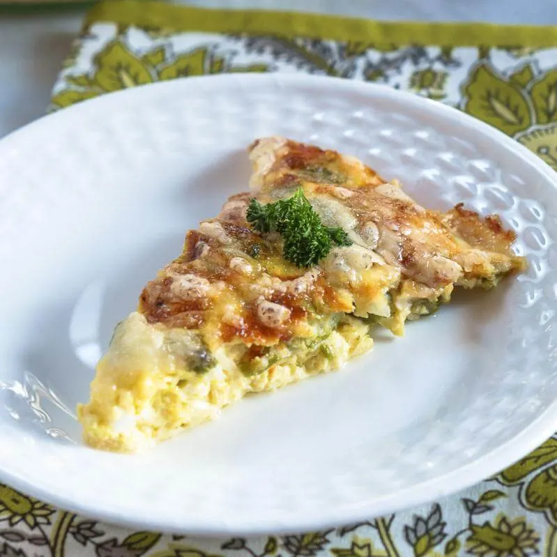 A slice of cheese asparagus quiche sitting on a white plate sitting over a green paisley napkin.