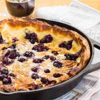 A cast-iron pan filled with a baked Blueberry Dutch Baby. A cup of coffee sits in the background.