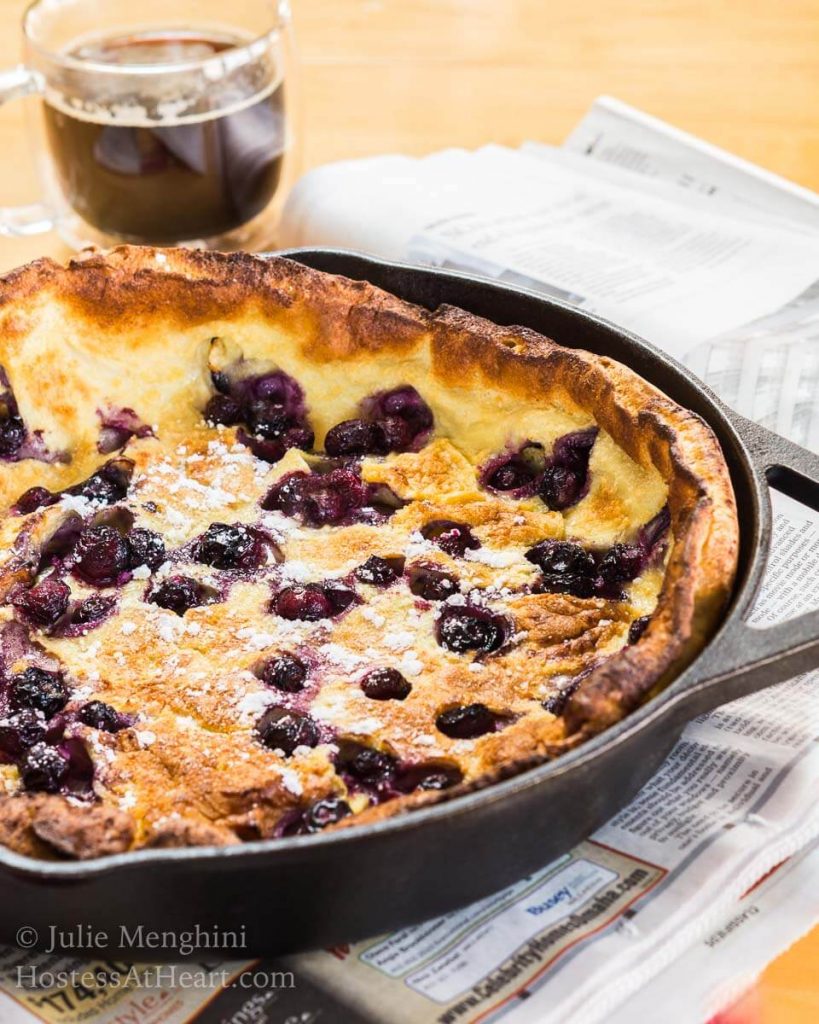 A blueberry Dutch Baby in a cast-iron pan sits over a newspaper with a cup of coffee sitting behind it.