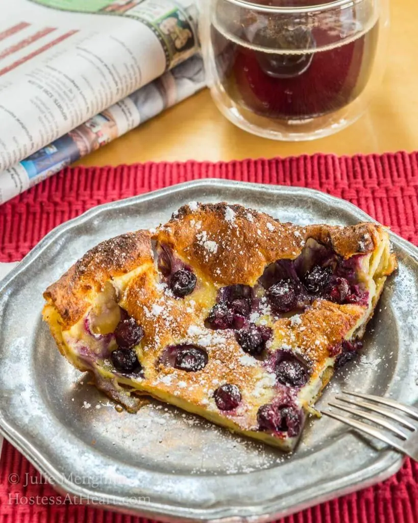 A slice of a blueberry dutch baby on a metal plate sits on a red placemat with a cup of coffee and a newspaper behind it.