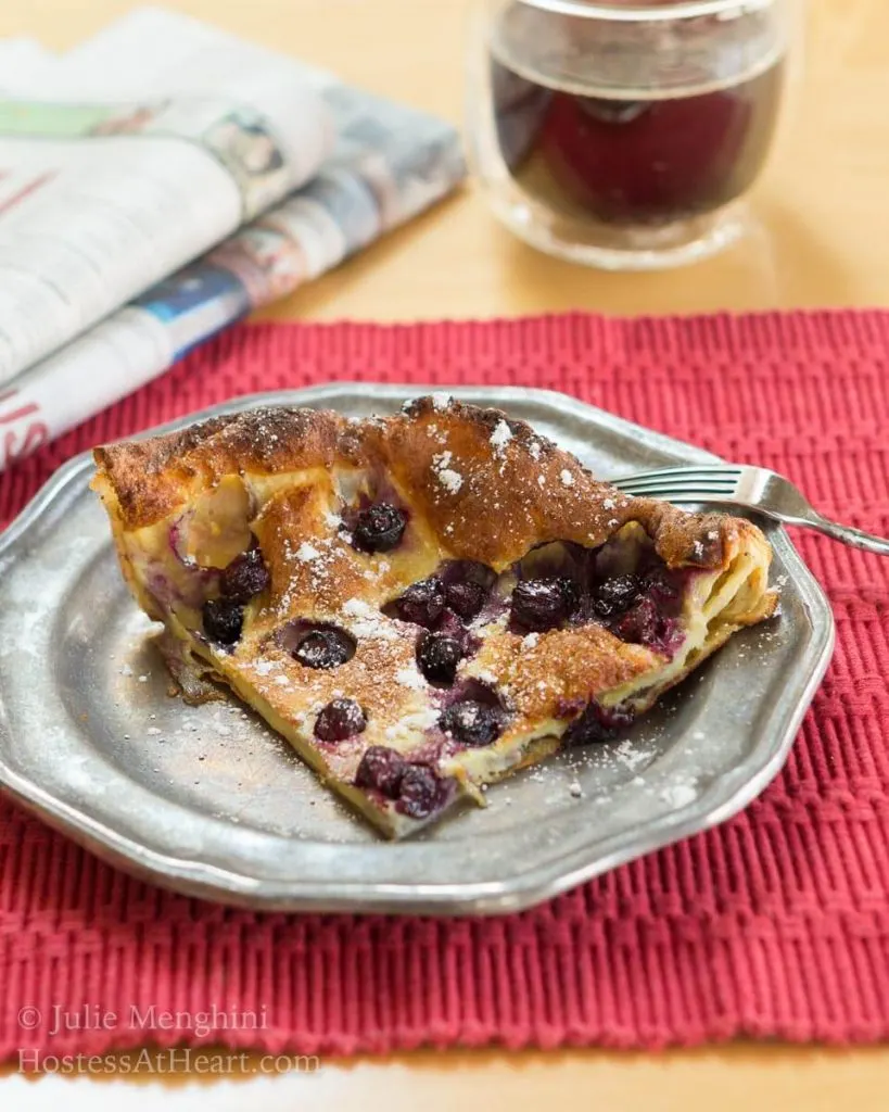 A slice of a Blueberry Dutch Baby pancake sitting on a metal plate over a red placemat. A cup of coffee and a newsletter sits in the background.