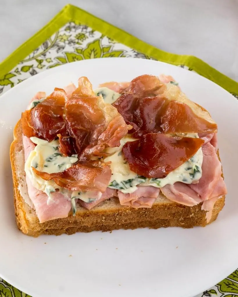 A slice of bread topped with sliced ham, brie, basil aioli, and crispy prosciutto.