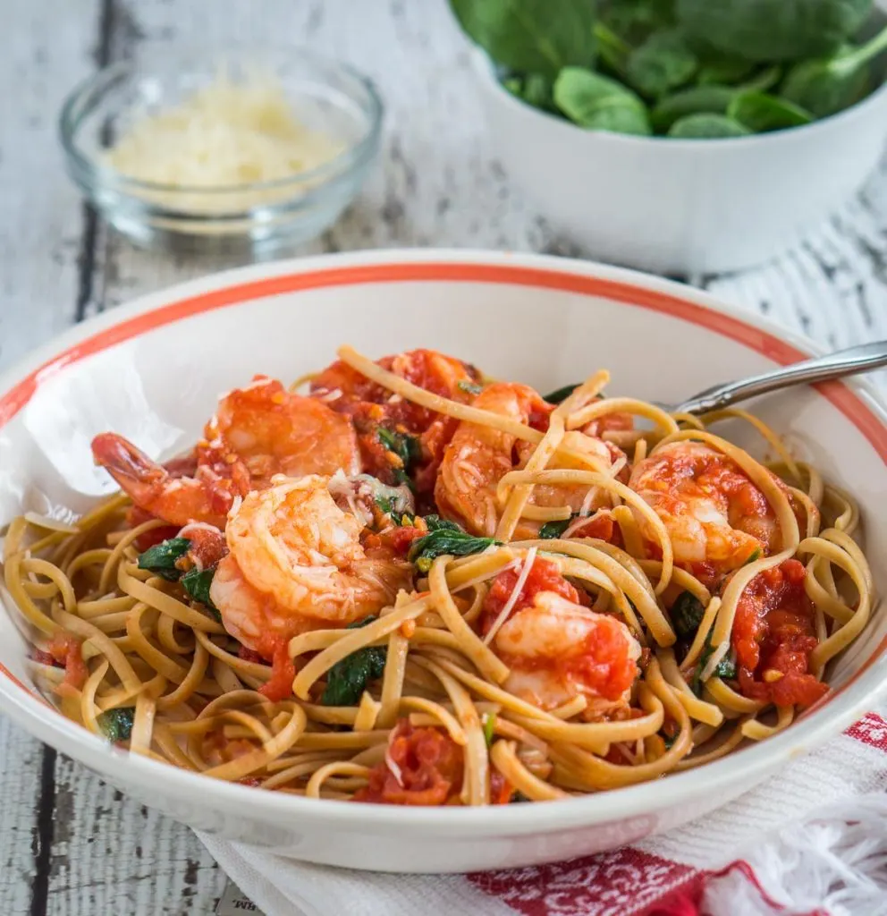 A white bowl with an orange stripe filled with fettuccine noodles in a tomato sauce topped with shrimp and sauteed spinach. A bowl of fresh spinach and a bowl of grated cheese sit in the background.