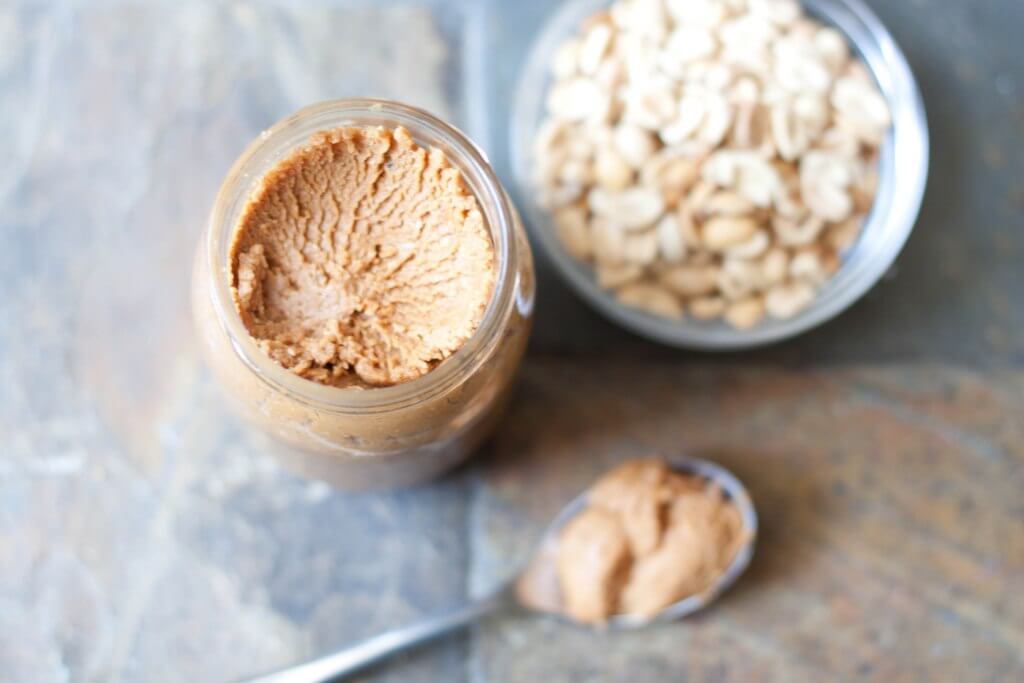 A jar of peanut butter made from scratch next to a bowl of raw peanuts.