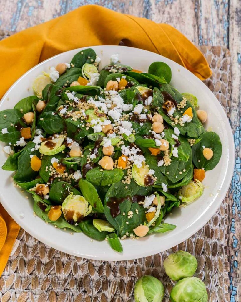 A white bowl filled with a spinach salad, shaved Brussels sprouts, garbanzo beans, and cheese sprinkled with balsamic vinaigrette.