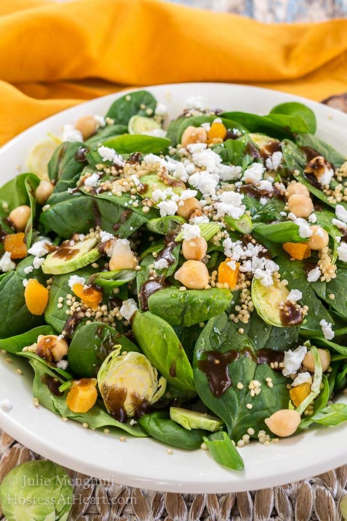 A close up of a white bowl filled with a salad, shaved Brussels sprouts, baby spinach, garbanzo beans, and cheese sprinkled with balsamic vinaigrette.