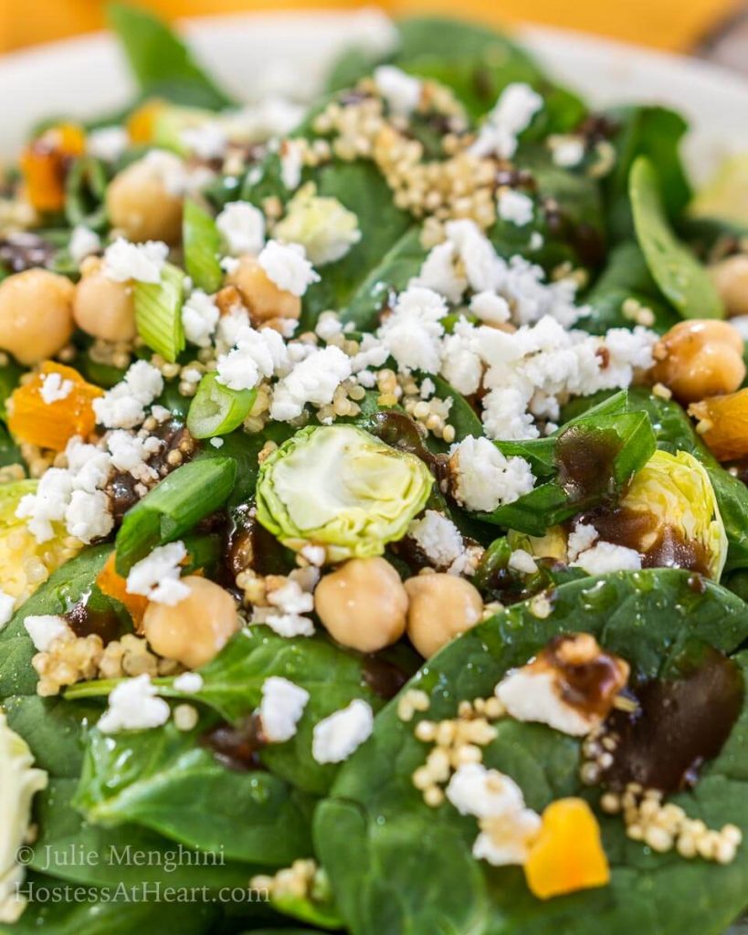 A close up of a white bowl filled with a salad consisting of shaved Brussels sprouts, spinach, garbanzo beans, and cheese sprinkled with balsamic vinaigrette.