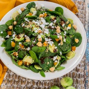 Top-down view of a salad made of spinach and shaved Brussels sprouts that's sprinkled with cheese and nuts. A gold napkin and whole Brussels sprouts sit on the sides over a wooden placemat.