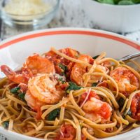 A white bowl with an orange stripe filled with fettuccine noodles in a tomato sauce topped with shrimp and sauteed spinach. A bowl of fresh spinach and a bowl of grated cheese sit in the background.