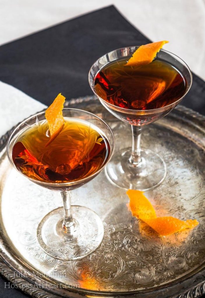 Two glasses of a whiskey cocktail sitting on a silver platter garnished with orange peels.