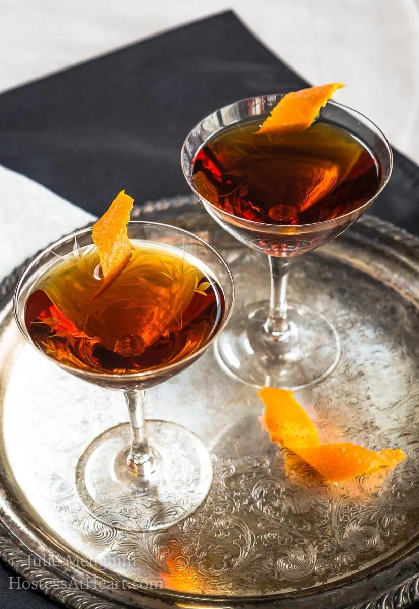 Two glasses of a whiskey cocktail sitting on a silver platter garnished with orange peels.